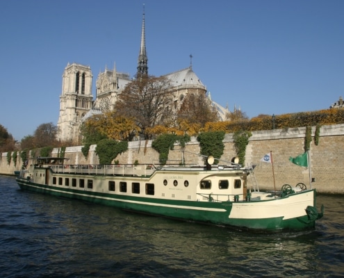 Paris by Boat - A cream and green canal boat sails on the Seine in Paris, with Notre Dame Cathedral and trees visible in the background.