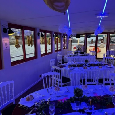 Paris en Bateau - Tables rondes dressées pour un événement formel avec des chaises blanches et des petits pains, décorées avec des ballons et de la verdure - idéal pour la privatisation de péniche pour différents événements, avec des fenêtres révélant de belles vues sur l'eau à l'extérieur.